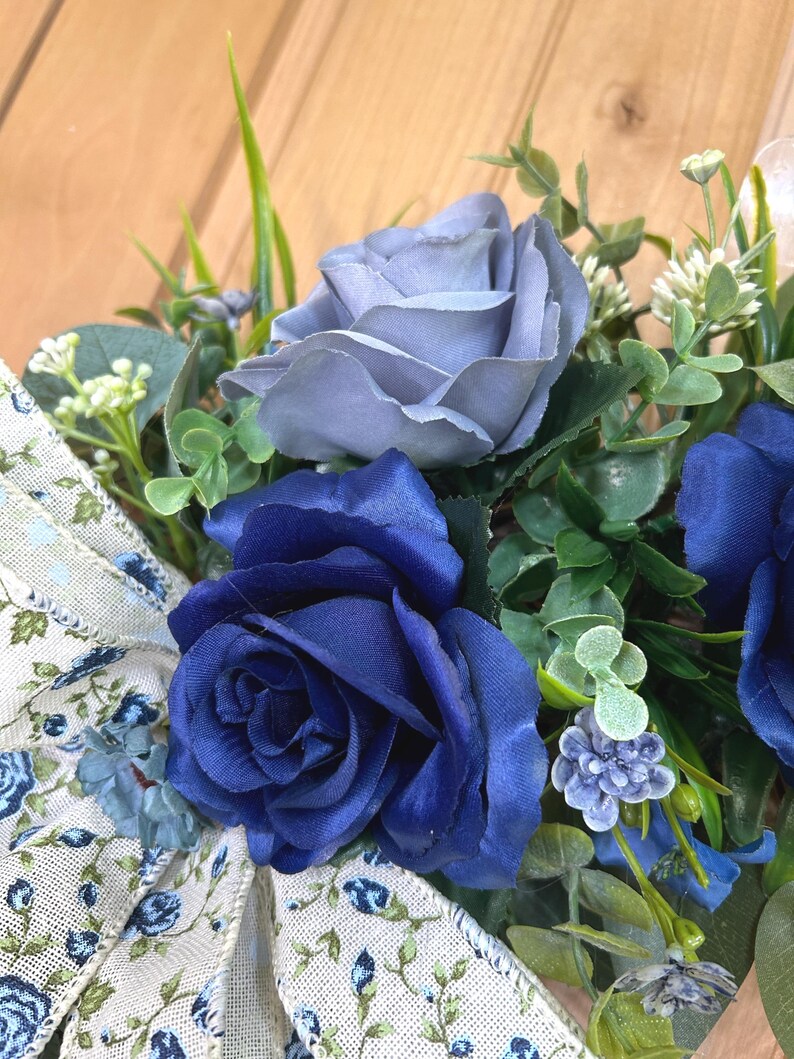 Op de afbeelding: Een bloemstuk met diep marineblauwe en stoffig blauwe rozen, geaccentueerd met groen blad, kleine witte bloemetjes en een beige lint met een blauw bloemenpatroon. De compositie bevat diverse bladeren en stengels tussen de rozen.
