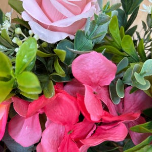 May include: A floral arrangement featuring a large, light pink rose as the focal point, surrounded by vibrant hot pink hydrangea petals and various green foliage, including small rounded leaves and sprigs of tiny white buds.