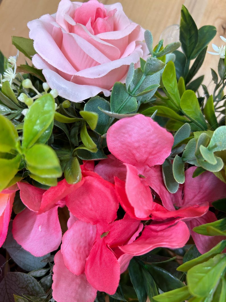 May include: A floral arrangement featuring a large, light pink rose as the focal point, surrounded by vibrant hot pink hydrangea petals and various green foliage, including small rounded leaves and sprigs of tiny white buds.