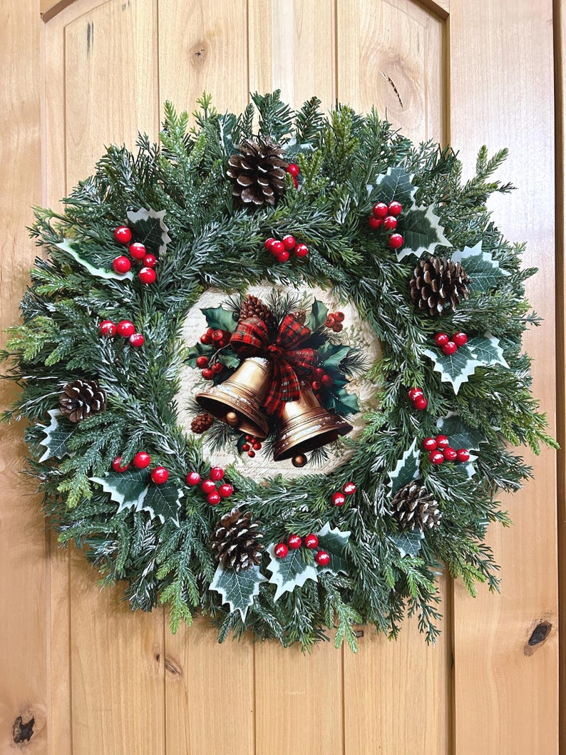 May include: A circular Christmas wreath featuring green pine needles, frosted tips, brown pinecones, and clusters of red berries. The center displays a round plaque with an illustration of two gold bells tied with a red and green plaid ribbon.