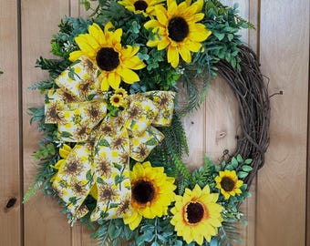 Op de afbeelding: Een ronde krans gemaakt van bruine takken, versierd met felgele zonnebloemen met donkerbruine harten. Groen blad en varens zijn ertussen verwerkt. Een grote decoratieve strik met een geel en wit ruitpatroon.