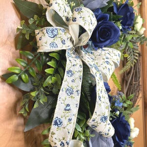 Op de afbeelding: Een decoratieve krans met kunstmatige donkerblauwe en lichtgrijze rozen, groen gebladerte en een cr&egrave;mekleurig lint met een blauw bloemenpatroon. Het arrangement is opgebouwd op een ronde basis van bruine takjes met diverse bladeren en witte bloemen.