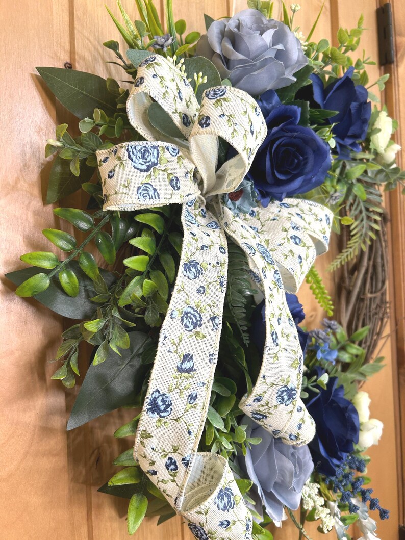 Op de afbeelding: Een decoratieve krans met kunstmatige donkerblauwe en lichtgrijze rozen, groen gebladerte en een cr&egrave;mekleurig lint met een blauw bloemenpatroon. Het arrangement is opgebouwd op een ronde basis van bruine takjes met diverse bladeren en witte bloemen.