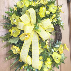 May include: A decorative wreath featuring bright yellow silk flowers, green fern leaves, and assorted foliage arranged on a circular brown twig base. A large, multi-looped yellow and white plaid ribbon is centered prominently on the floral arrangement.