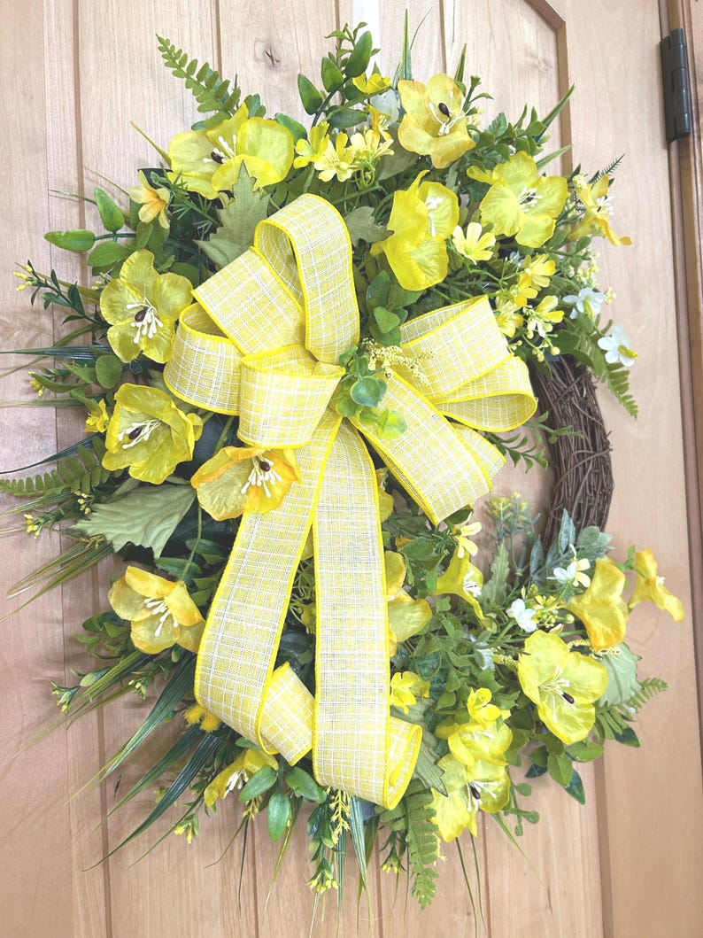 May include: A decorative wreath featuring bright yellow silk flowers, green fern leaves, and assorted foliage arranged on a circular brown twig base. A large, multi-looped yellow and white plaid ribbon is centered prominently on the floral arrangement.