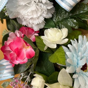 May include: A decorative floral arrangement featuring a large white carnation, vibrant pink hydrangea blossoms, a light blue daisy, and smaller white roses. The arrangement is accented with green leaves and a light blue and white striped fabric ribbon.