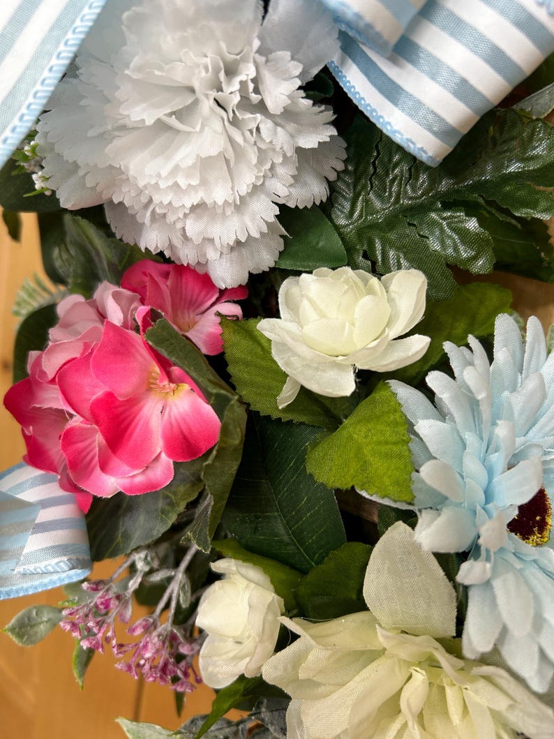 May include: A decorative floral arrangement featuring a large white carnation, vibrant pink hydrangea blossoms, a light blue daisy, and smaller white roses. The arrangement is accented with green leaves and a light blue and white striped fabric ribbon.