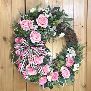 May include: A round wreath constructed from a brown twig base, adorned with artificial pink roses, white blossoms, and green foliage. A decorative bow featuring a black and white striped pattern with a pink floral print is attached to the side.