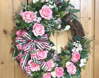 May include: A round wreath constructed from a brown twig base, adorned with artificial pink roses, white blossoms, and green foliage. A decorative bow featuring a black and white striped pattern with a pink floral print is attached to the side.