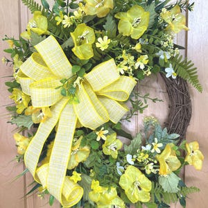 May include: A circular wreath featuring a brown twig base adorned with bright yellow silk flowers, various green foliage, and fern fronds. A large, multi-looped yellow and white checkered ribbon bow is attached to the left side of the arrangement.