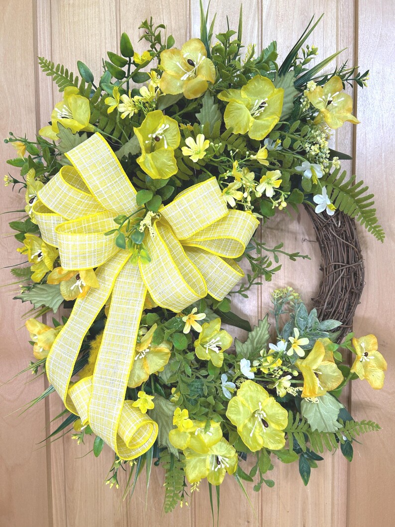 May include: A circular wreath featuring a brown twig base adorned with bright yellow silk flowers, various green foliage, and fern fronds. A large, multi-looped yellow and white checkered ribbon bow is attached to the left side of the arrangement.