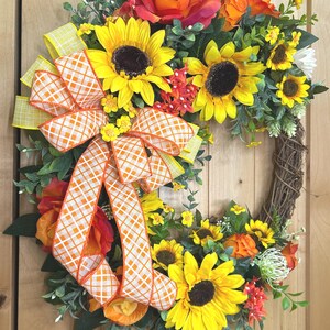 May include: A circular wreath featuring yellow sunflowers, orange roses, and green foliage on a brown twig base. A large decorative bow made of orange and white checkered ribbon with yellow edges is attached to the left side of the floral arrangement.