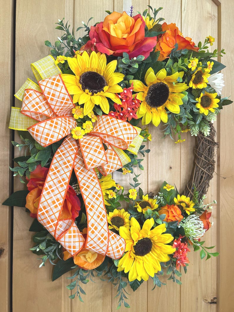 May include: A circular wreath featuring yellow sunflowers, orange roses, and green foliage on a brown twig base. A large decorative bow made of orange and white checkered ribbon with yellow edges is attached to the left side of the floral arrangement.