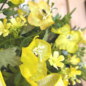 May include: A decorative wreath featuring bright yellow silk flowers with prominent white stamens, interspersed with green foliage and sprigs. A yellow and white checkered ribbon is tied at the base, adding a structured pattern to the floral arrangement.