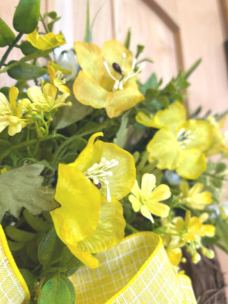 May include: A decorative wreath featuring bright yellow silk flowers with prominent white stamens, interspersed with green foliage and sprigs. A yellow and white checkered ribbon is tied at the base, adding a structured pattern to the floral arrangement.