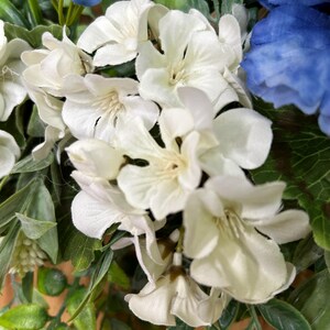 May include: A floral arrangement featuring clusters of white blossoms and deep blue flowers interspersed with green leaves. The white petals have a soft, layered appearance, while the blue flowers provide a vibrant contrast against the foliage.