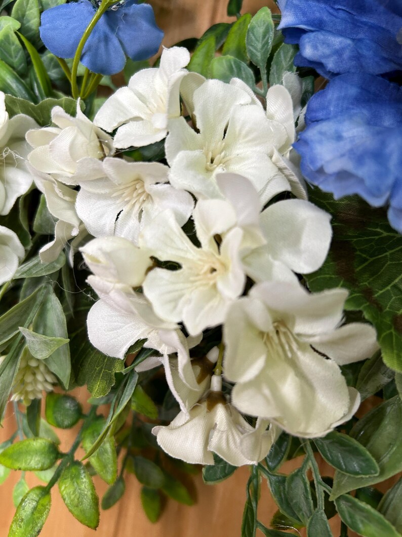 May include: A floral arrangement featuring clusters of white blossoms and deep blue flowers interspersed with green leaves. The white petals have a soft, layered appearance, while the blue flowers provide a vibrant contrast against the foliage.