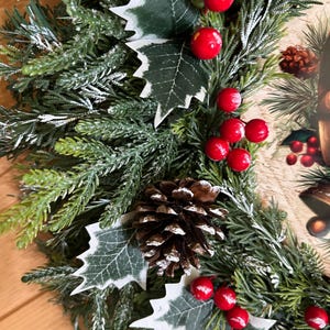 May include: A holiday wreath featuring artificial green pine needles with a light dusting of white snow. The wreath is decorated with clusters of bright red berries, dark green holly leaves with white edges, and a brown pinecone in the center.