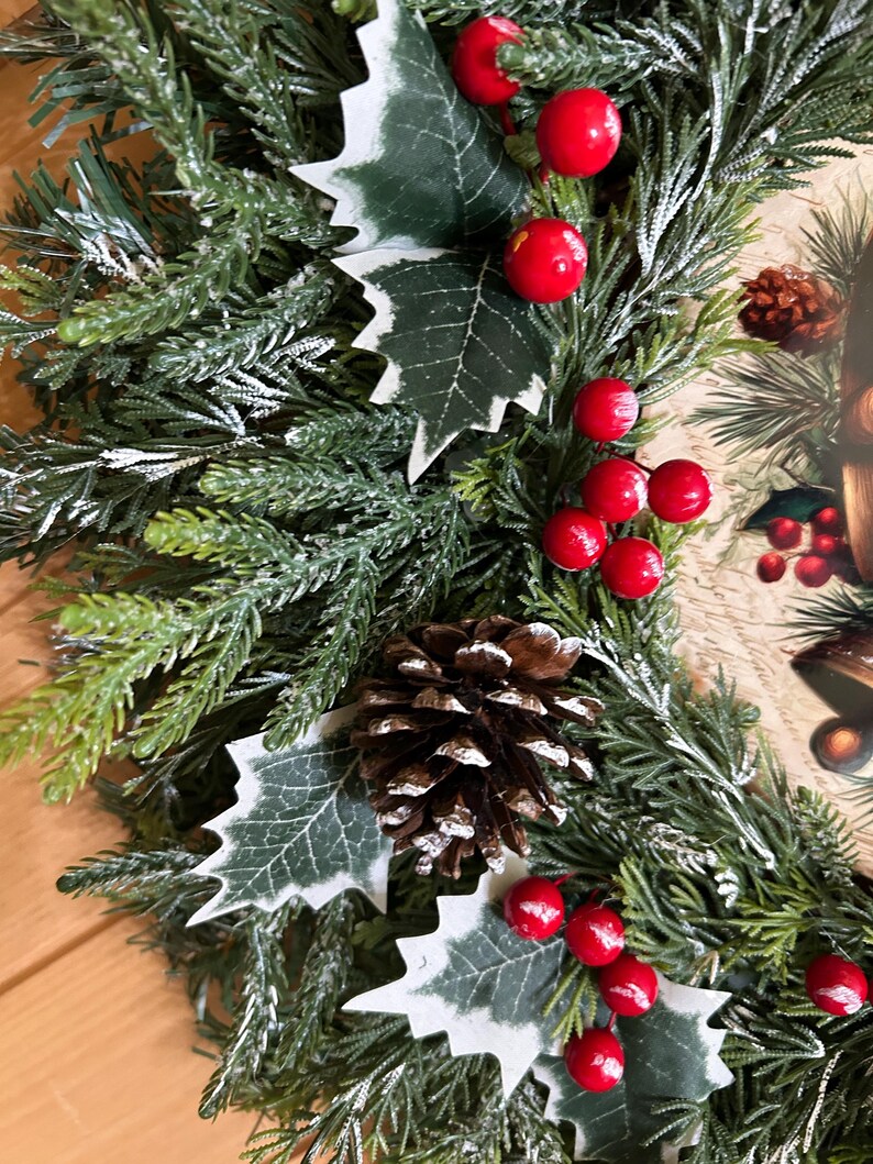 May include: A holiday wreath featuring artificial green pine needles with a light dusting of white snow. The wreath is decorated with clusters of bright red berries, dark green holly leaves with white edges, and a brown pinecone in the center.
