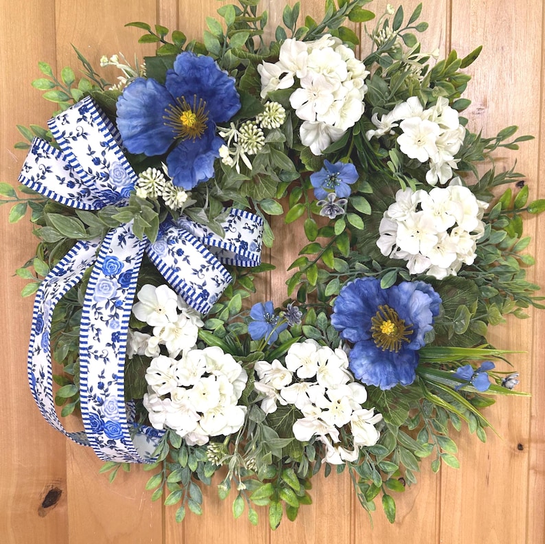 May include: A circular wreath featuring large deep blue flowers with yellow centers, clusters of white hydrangea-like blooms, and green foliage. A decorative ribbon with a blue and white floral pattern and striped edges is tied to the side.