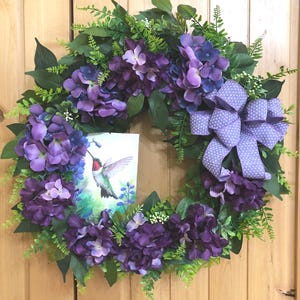 May include: A decorative wreath featuring clusters of purple and blue hydrangea flowers, green fern foliage, and a light purple polka dot ribbon bow. A rectangular sign depicting a hummingbird in flight is nestled among the floral arrangement.