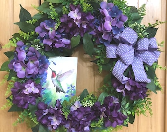 May include: A decorative wreath featuring clusters of purple and blue hydrangea flowers, green fern foliage, and a light purple polka dot ribbon bow. A rectangular sign depicting a hummingbird in flight is nestled among the floral arrangement.