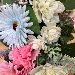 May include: A floral arrangement featuring light blue gerbera daisies, vibrant pink carnations, and creamy white blossoms. The bouquet includes various green leaves, small white filler flowers, and textured fern-like foliage, creating a dense display.