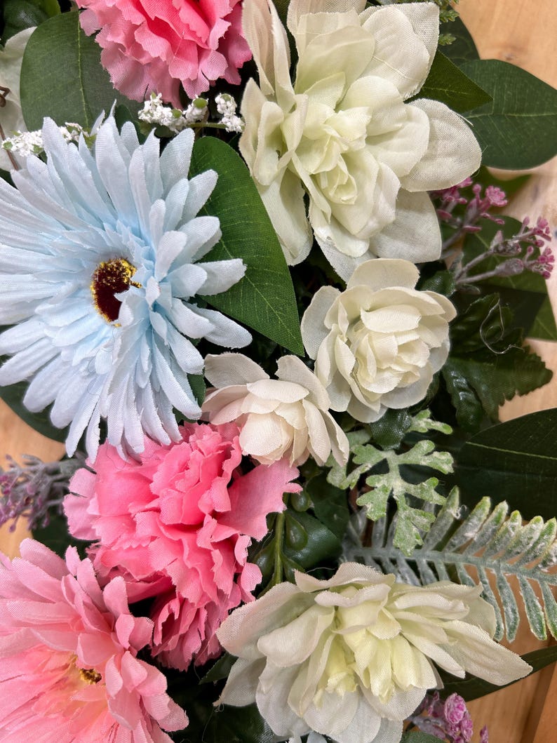 May include: A floral arrangement featuring light blue gerbera daisies, vibrant pink carnations, and creamy white blossoms. The bouquet includes various green leaves, small white filler flowers, and textured fern-like foliage, creating a dense display.