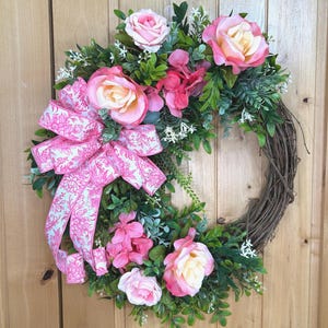 May include: A circular wreath featuring a natural brown twig base adorned with artificial pink and cream roses, green foliage, and small white flowers. A large decorative ribbon with a pink and white floral pattern is attached to the side.