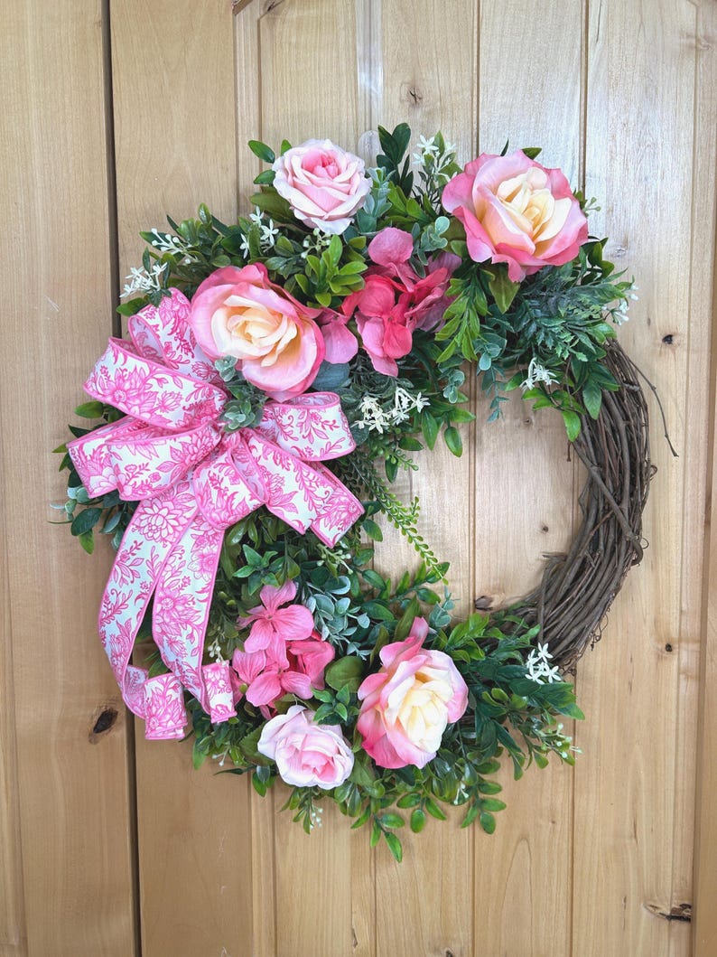 May include: A circular wreath featuring a natural brown twig base adorned with artificial pink and cream roses, green foliage, and small white flowers. A large decorative ribbon with a pink and white floral pattern is attached to the side.