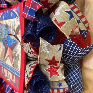 May include: A patriotic wreath featuring red, white, and blue mesh ribbons, burlap ribbons with red glitter stars, and a rectangular sign with a star graphic and the word AMERICA. The design incorporates plaid, striped, and solid coloured fabric.
