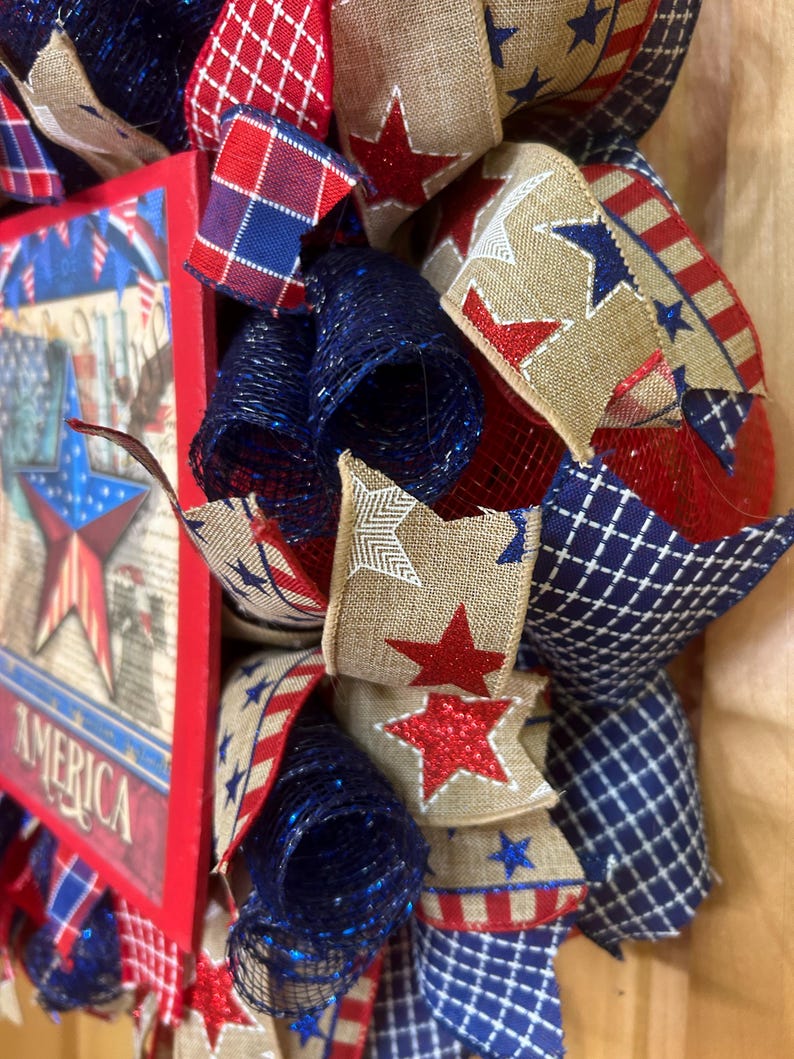 May include: A patriotic wreath featuring red, white, and blue mesh ribbons, burlap ribbons with red glitter stars, and a rectangular sign with a star graphic and the word AMERICA. The design incorporates plaid, striped, and solid coloured fabric.