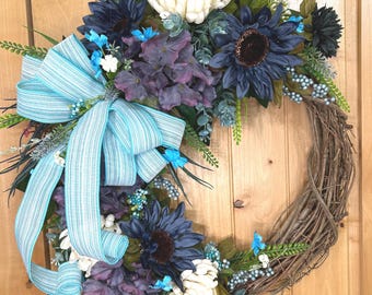 May include: A decorative wreath featuring dark blue sunflowers, cream-colored dahlias, and purple hydrangea blossoms. The arrangement includes green foliage, small light blue berries, and a large, multi-looped light blue and white striped ribbon.