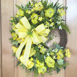 May include: A circular wreath featuring a brown twig base adorned with bright yellow flowers, various green foliage, and long grass-like stems. A large, multi-looped yellow and white plaid ribbon is attached to the left side of the floral arrangement.
