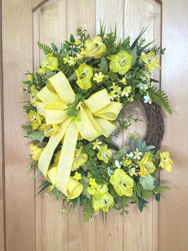 May include: A circular wreath featuring a brown twig base adorned with bright yellow flowers, various green foliage, and long grass-like stems. A large, multi-looped yellow and white plaid ribbon is attached to the left side of the floral arrangement.