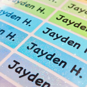 May include: Close-up of a set of blue and green personalised name labels with glitter. The labels read "Jayden H."