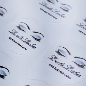 May include: Close-up of several white round stickers with a stylized illustration of a closed eye with eyelashes. The text "Loveli Lashes" and "100% Real Mink Lashes" is printed on each sticker.