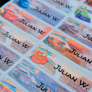May include: A sheet of colorful, glittery name labels with the name "Julian W." printed on each label. The labels feature images of cars from the Disney Pixar movie "Cars".