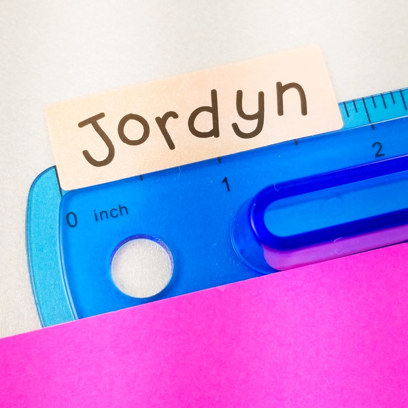 Op de afbeelding: Een roze plakbriefje met de naam "Jordyn" erop geschreven in zwarte stift. Het plakbriefje ligt op een blauwe plastic liniaal met een witte "0 inch" markering.