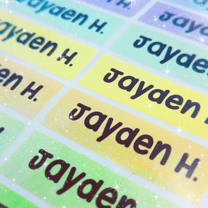 May include: A close-up of colourful, glittery name labels with the name "Jayden" printed on them. The labels are in various shades of green, yellow, and blue.