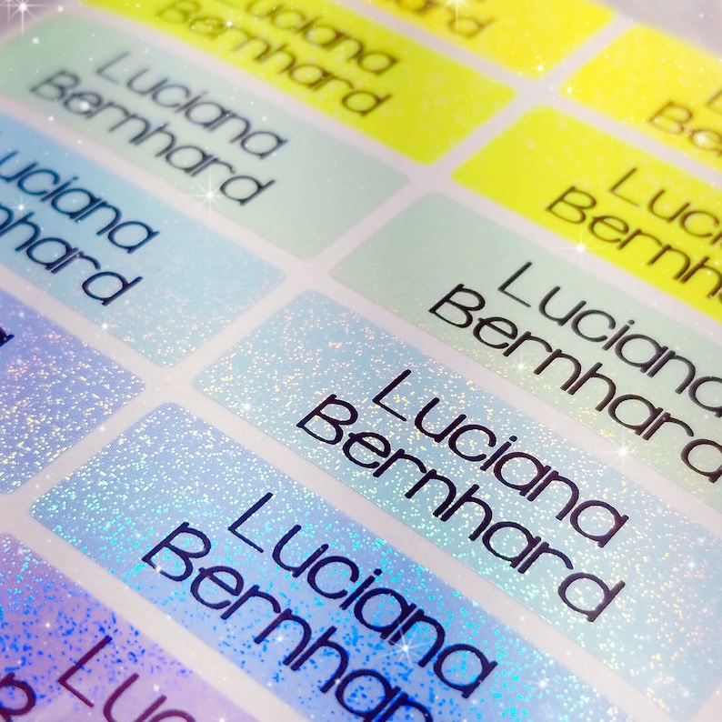 Op de afbeelding: Een close-up van iriserende blauwe en witte glitter-naamstickers met de naam "Luciana Bernhard" in zwart gedrukt.