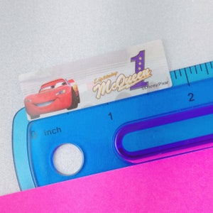 May include: A red race car with a smiling face is on a sticker with the text "Lightning McQueen" and a number "1" with stars. The sticker is on top of a blue ruler with inch markings.