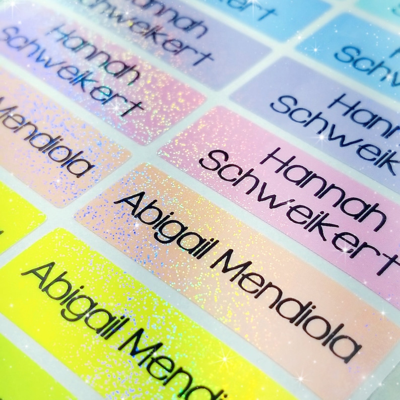 Op de afbeelding: Close-up van kleurrijke, glinsterende naamstickers met de namen "Hannah Schweikert" en "Abigail Mendiola" in het zwart gedrukt.