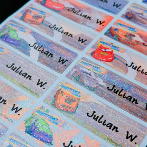 May include: A sheet of colorful, glittery, holographic name labels featuring a Cars theme. The labels have the text "Julian W." and a picture of a red race car with the words "Drifting Time" on the side.