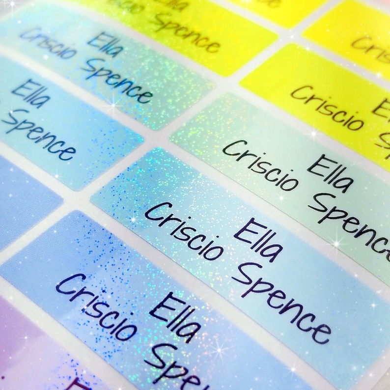 Op de afbeelding: Close-up van blauwe en witte, iriserende glitter-naamstickers met de tekst "Criscio Ella Spence" die op elke sticker wordt herhaald.