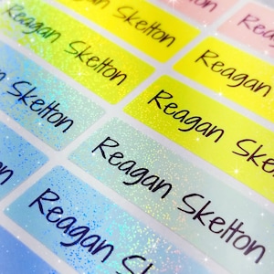 Op de afbeelding: Een close-up van kleurrijke, glinsterende naamstickers met de naam "Reagan Skelton" erop. De stickers zijn perfect voor schoolspullen, lunchboxen of andere persoonlijke items.