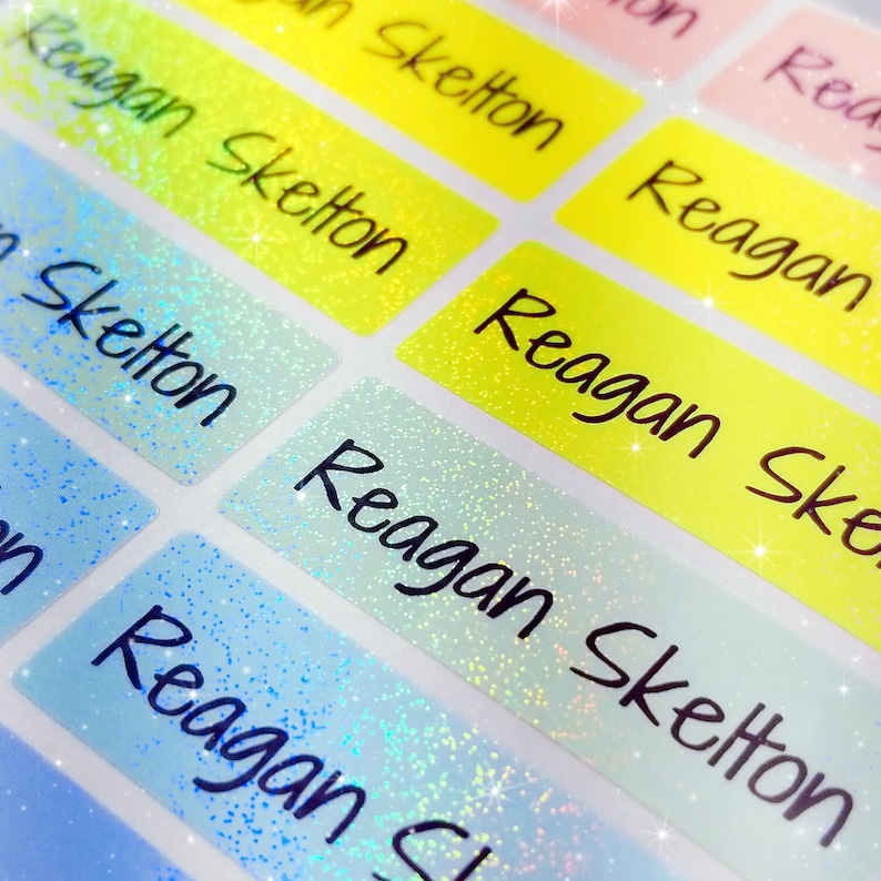 Op de afbeelding: Een close-up van kleurrijke, glinsterende naamstickers met de naam "Reagan Skelton" erop. De stickers zijn perfect voor schoolspullen, lunchboxen of andere persoonlijke items.