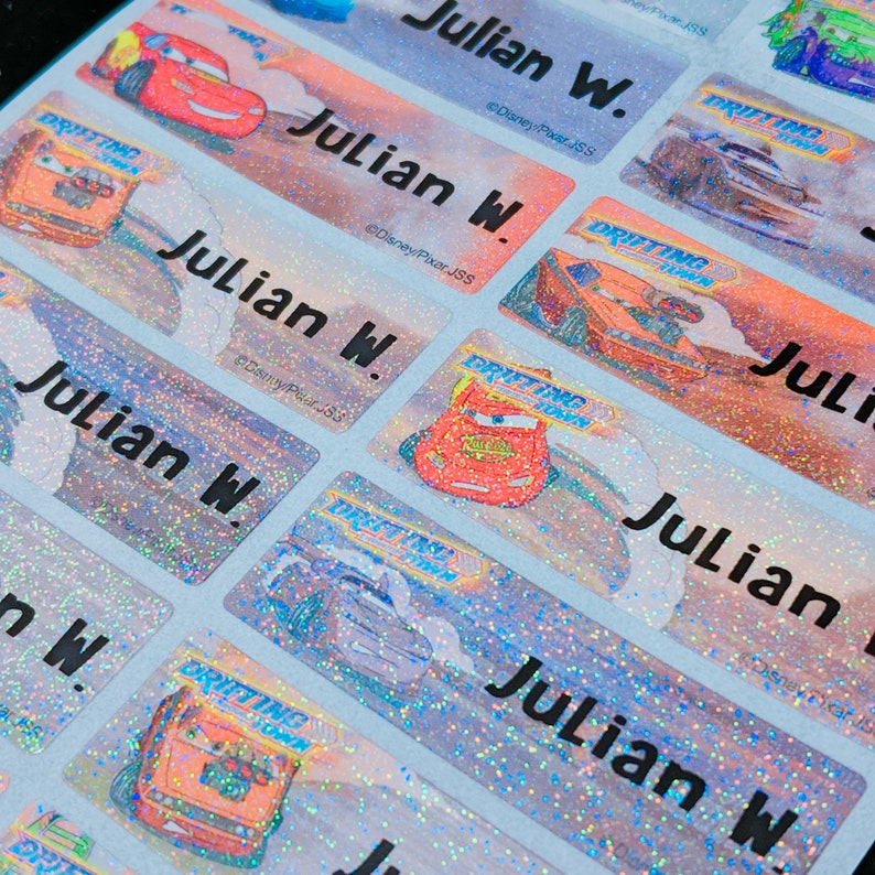 May include: A close-up of several colorful, glittery name labels featuring a cartoon car racing. The labels are printed with the name "Julian W." and the text "Disney/Pixar JSS".