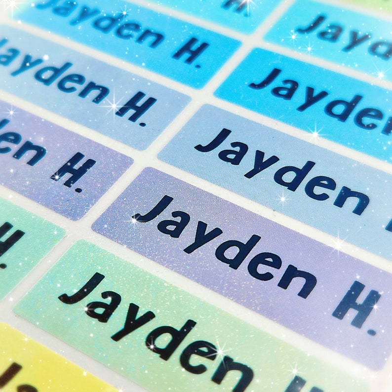 May include: Close-up of colourful, glittery name labels with the name "Jayden H." printed in black. The labels are rectangular and have a holographic finish.