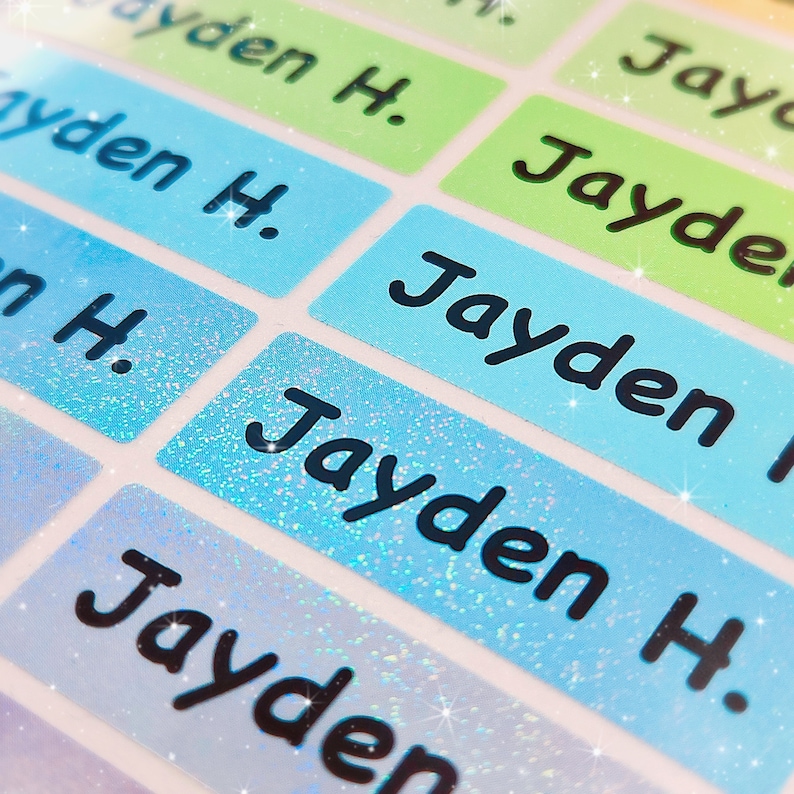 May include: Close-up of a set of blue and green personalised name labels with glitter. The labels read "Jayden H."